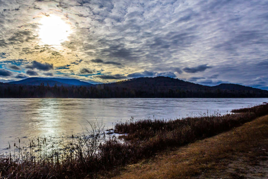 a mountain landscape that includes a body of water in the foreground and a cloudy sky with a bright sun shining in the background