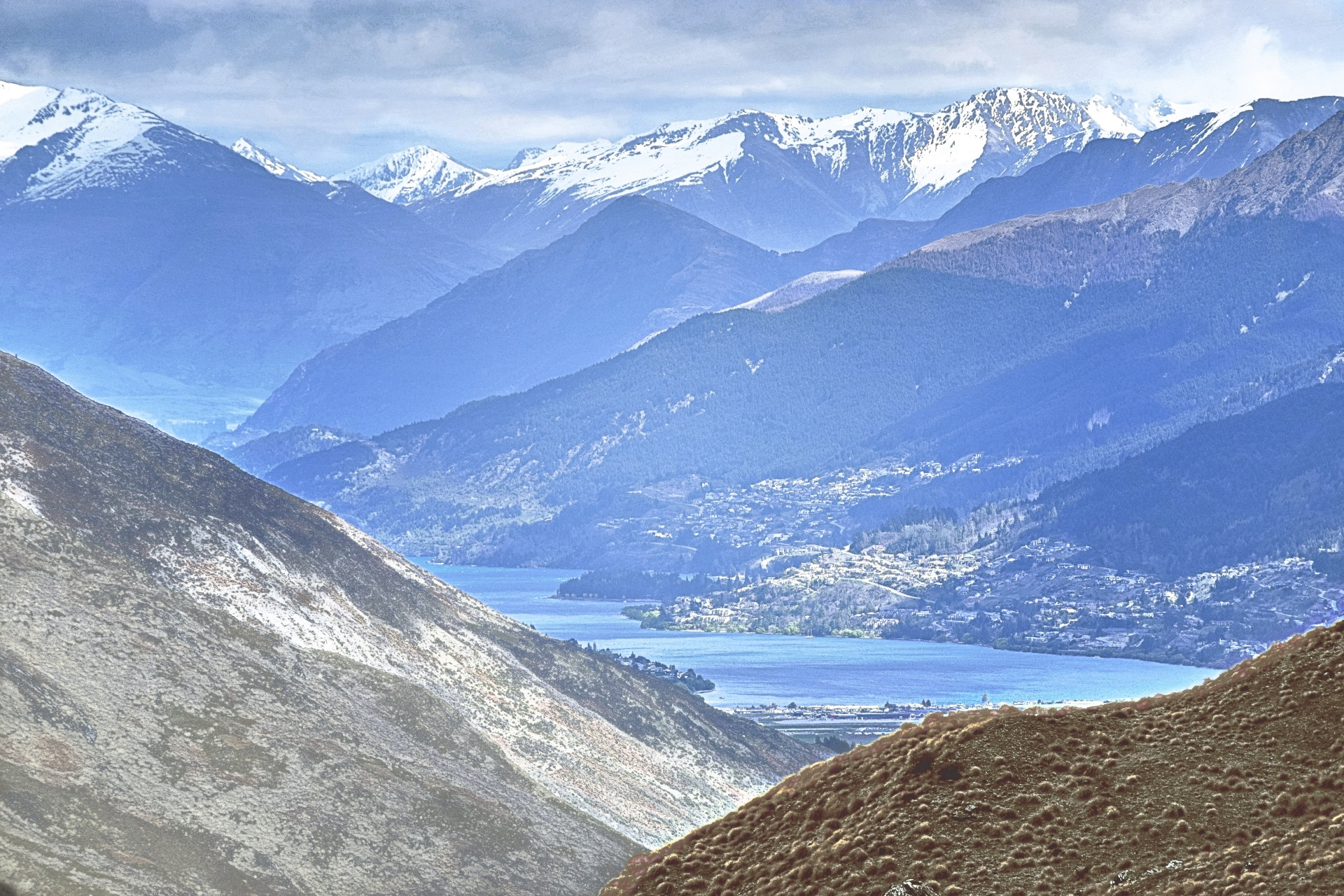 a snowy mountain landscape with a gray, cloudy sky and a small body of water in the foreground, mostly hidden by more mountains