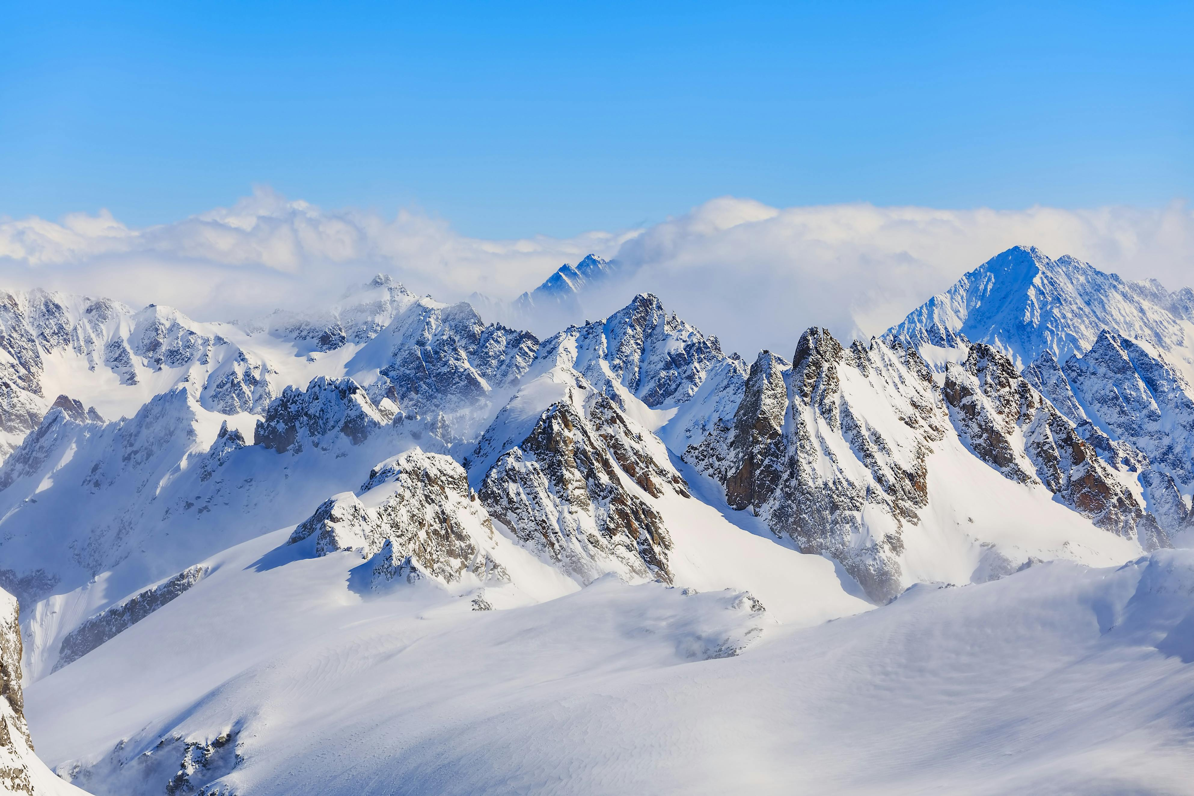 a dramatic mountain landscape with lots of snow; the background is cloudy near the mountains, and there is clear blue sky above the clouds